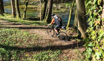 Ein Radfahrer mit einem Rucksack fährt auf einem schmalen Waldweg neben einem Fluss, umgeben von Bäumen und grünem Unterholz, bei sonnigem Wetter.